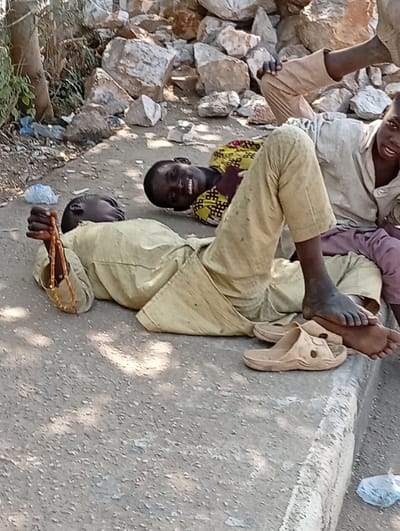 Three children are reclining on a roadside. One child is lying down, holding a string of beads. Adjacent to this child is another, grinning at the camera. A third child is positioned higher up in the scene. Discarded plastic bags litter the ground. The scene is set alongside a concrete road and a pile of rocks. One pair of sandals are laying on the ground next to the reclining child.
