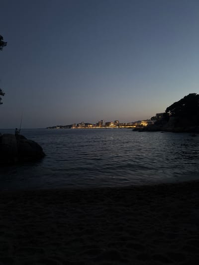 The image depicts a coastal scene at dusk in Castell d'Aro, Platja d'Aro i s'Agaró, Spain. A lone figure, silhouetted against the darkening sky, is fishing from a cluster of large rocks on the left side of the frame. The sea, appearing dark and rippled, occupies the middle ground. In the distance, a city skyline is illuminated with numerous lights, suggesting a developed urban area along the coast. The foreground consists of dark, textured sand, presumably a beach. A line of trees and rocky outcrops is visible along the coastline to the right, silhouetted against the twilight sky. The overall atmosphere is serene and captures the transition from daylight to night.