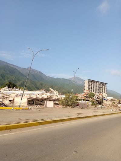 The image depicts the aftermath of a catastrophic event in Caraballeda, Venezuela, with two partially collapsed buildings as the central focus. The building on the left is almost entirely reduced to rubble, with large concrete slabs and debris scattered across the site. Some sections of the lower floors are still discernible, showing concrete walls and exposed rebar. A leaning lamppost stands in front of this wreckage.

To the right, another building, a multi-story structure, has also suffered severe damage. While parts of its facade remain intact, large sections have collapsed, creating a chaotic jumble of concrete, metal, and building materials. The upper floors of this structure appear somewhat more stable, but the overall impression is one of significant structural compromise. Several lampposts are visible, some of which are also damaged.

The surrounding environment shows signs of destruction, with debris extending towards the foreground. A road with a yellow curb runs in front of the damaged buildings. In the background, lush green mountains rise under a clear blue sky. The scene is devoid of any visible people or ongoing activity, suggesting the image was taken sometime after the event, likely for documentation purposes. The overall atmosphere is one of desolation and destruction, highlighting the impact of a severe disaster.
