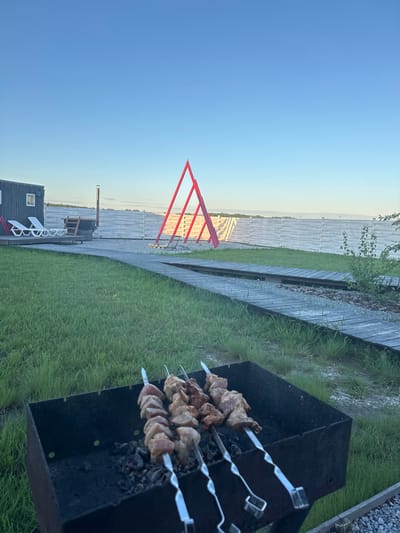 The image presents an outdoor scene, likely in a residential area, given the presence of a house with white outdoor furniture and a fence. A barbecue grill is positioned on the grass, with skewers containing meat cooking over charcoal. In the background, there's a bright red triangular structure standing prominently in the landscape. The sky is a clear blue, suggesting fair weather. Based on the request, it is assumed this is located in the city of Pad', Russia.