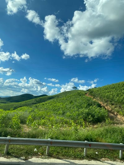 The image captures a verdant landscape with rolling hills covered in lush green vegetation. The sky above is a vibrant blue, dotted with fluffy white clouds. A dirt path winds its way up one of the hills, suggesting a rural or undeveloped area. In the foreground, a silver guardrail runs parallel to a paved surface, possibly a road. The scene is set in Thôn 18, Vietnam, and it appears to be a bright, sunny day. There are no people or animals visible in the image. The environment is natural and tranquil, with the focus on the expansive landscape and the interplay of light and shadow on the hills.