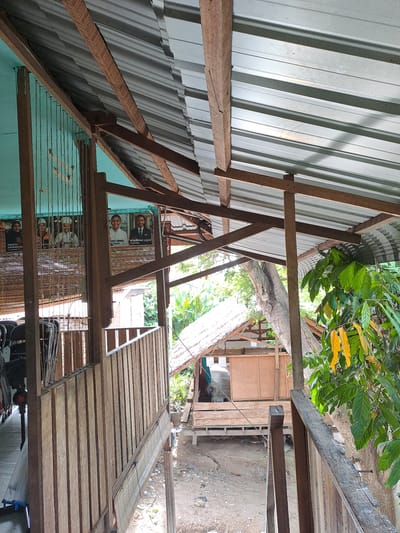 The image depicts a rustic, open-air structure with a corrugated metal roof supported by wooden beams, located in Geulanggang Baro, Indonesia. The scene suggests a partly built or maintained area. In the background, a small wooden building with a thatched roof is visible amidst lush green foliage and a large tree. Several framed portraits, seemingly of individuals in formal attire, are displayed on a light blue wall. Wooden railings and a pathway made of wooden planks are prominent features, indicating a raised walkway or balcony. The ground below appears to be unpaved. The overall impression is one of a rural or semi-rural setting, possibly a community center or a residential area with open-air extensions.