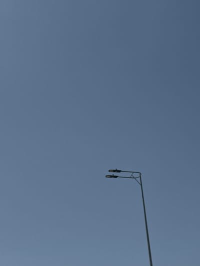 A low-angle shot shows an outdoor lighting fixture against a clear, overcast sky. The fixture is mounted on a metal pole extending horizontally from the upper right of the frame. The pole has a short vertical section that terminates in two downward-facing lights. The location is likely Hof Ashkelon Regional Council, Israel.