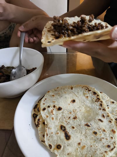 In Imus, Philippines, a close-up shot captures a person's hands holding a piece of flatbread filled with a ground meat mixture. Another hand is seen using a spoon to serve the meat from a white bowl. In the foreground, a white plate displays several pieces of lightly browned flatbread. The scene appears to be set at a table with a wooden surface.