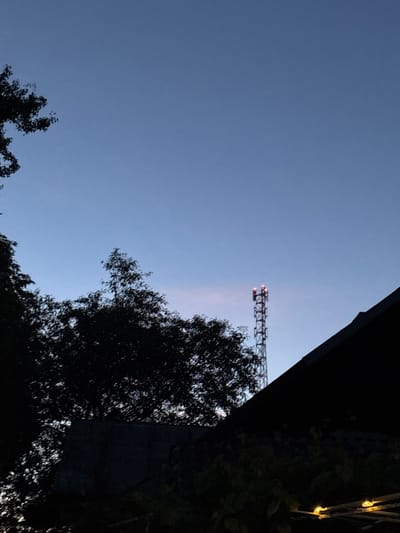The image depicts a twilight scene with a clear, blue sky. In the background, a tall, lattice-style communication tower stands prominently, illuminated by two red lights at its apex. To the left, the silhouette of a dense tree fills a significant portion of the frame. In the foreground, the dark, angular roofline of a building dominates the lower right side, with hints of foliage and some small, glowing yellow lights visible beneath it. The overall atmosphere suggests an outdoor setting during the evening, possibly in a semi-rural or suburban area within the Ганчарскі сельскі Савет, Belarus, given the presence of both natural elements like trees and man-made structures like the tower and building. No people are visible in the image.