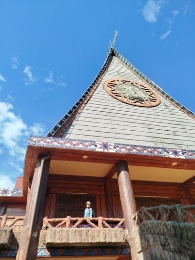 A low-angle, eye-level shot reveals a large wooden building under a bright blue sky with scattered clouds. The building's roof is steeply pitched and triangular, with a circular emblem near the peak. The emblem appears to be a carving or relief. The building's design incorporates intricate geometric patterns along the eaves. Below the roof, a balcony with a textured, faux-stone railing is visible. A single person wearing a hat and backpack stands on the balcony. The building's architectural style suggests a cultural or religious function. The image was taken in An Phu Ward, Vietnam.