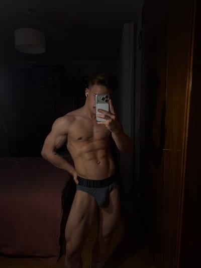 A muscular man in gray briefs poses in a mirror selfie. His phone obscures his face, but he wears an earbud in his left ear. His torso shows defined abs and a tattoo is visible on his chest. A bed with a dark red pillow is partially visible in the lower right corner of the frame. A dark wooden dresser is above him, and a sconce is seen to his left.