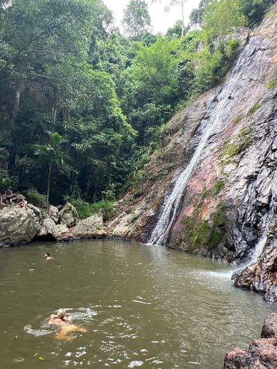 The image shows a natural waterfall with lush green vegetation on the surrounding cliffs. In the foreground, a large pool of murky water is visible, with two individuals swimming. One person, with blonde hair, is swimming closer to the viewer, creating ripples in the water. Another person, with dark hair, is swimming further away. Several large rocks are scattered around the edge of the pool, and some people are sitting on them in the background. The waterfall itself cascades down a rocky, wet cliff face, with water spraying at the bottom into the pool. The environment suggests a tropical or subtropical climate.