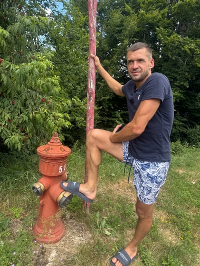 A man stands outdoors, leaning on a red-painted wooden pole. He is wearing a dark blue t-shirt with a logo on the left chest and white and blue patterned shorts. His right leg is bent, with his foot resting on a red fire hydrant. The fire hydrant has the number "58" painted in white on its side. The man is looking towards the camera.
The environment is grassy with several green trees in the background. There are also some leafy plants with red berries visible to the left of the fire hydrant. The scene appears to be in a rural or semi-rural area, possibly a park or roadside. The weather is overcast, as indicated by the diffused lighting and the sky visible through the trees. There is no specific information in the image to confirm the location in Les Rousses, France.