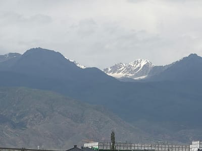 The image depicts a mountainous landscape with snow-capped peaks in the background, possibly part of the Tian Shan mountain range. In the foreground, there are hills with sparse vegetation. A partially constructed building with scaffolding is visible at the bottom of the image, suggesting ongoing development or renovation. The sky is overcast, indicating a cloudy day. The scene appears to be a rural or semi-urban area within the city of Kara-Oy, Kyrgyzstan, characterized by its natural beauty and developing infrastructure. There are no people visible in the image.