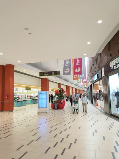 The image shows the interior of a shopping mall, possibly in Cebalat, Tunisia, with a wide tiled corridor. On the left, there are orange structural columns and a supermarket with checkout counters and shelves visible in the background. A digital screen with "digicom" logo is present. Further down the corridor, there are colorful banners hanging from the ceiling with various icons and text. On the right, there are shop fronts with mannequins displayed in windows, including one labeled "OXFORD CLUB". Several people are walking in the corridor, some with shopping carts. The lighting is bright, suggesting an indoor environment.