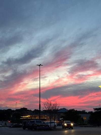 The image depicts a vibrant, colorful sunset in a location near Oak Brook, Illinois, United States. The sky is filled with streaks of pink, white, and gray clouds, indicating either sunrise or sunset. In the foreground, a tall streetlight with a bird deterrent is visible. Beyond the streetlight, there is a parking area adjacent to a road. Vehicles including cars and SUVs are parked and driving, indicating a commercial or residential area. Trees are also visible. The overall lighting suggests a time near dusk.