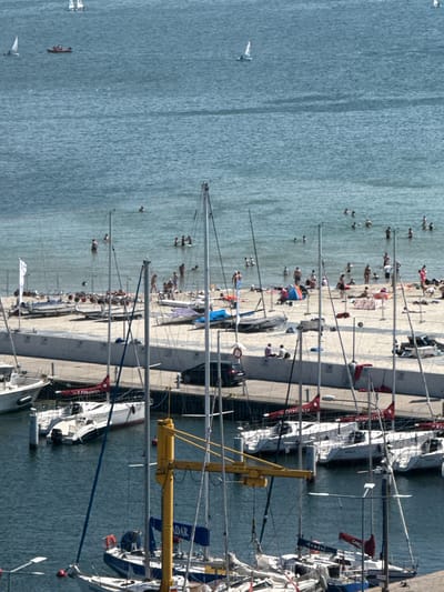The image depicts a lively scene at a harbor in Gdynia, Poland, likely during a sunny day. The water, a calm blue, is dotted with several sailboats docked along a quay. People are scattered on a sandy beach and in the water, enjoying the weather. Some are wading or swimming, while others are sunbathing on the shore or walking along the waterfront.

The boats are a prominent feature, with their tall masts creating a striking silhouette against the sky. One sailboat has red sails with white lettering that appears to read "GDYNIA" and possibly "POLISH". Another boat has a yellow structure attached to its mast. The quay itself is a concrete walkway lined with more sailboats and various objects, including what might be beach chairs and umbrellas.

Visible text on the boats includes "POLISH" and possibly "YACHT CLUB" on one sail, and "ADAR" and "CAROLINA" on others.

The overall atmosphere seems relaxed and recreational, with people enjoying a day by the sea. The presence of numerous sailboats suggests this is a popular spot for sailing or a maritime event. The clear sky and bright sunlight indicate good weather conditions for outdoor activities.