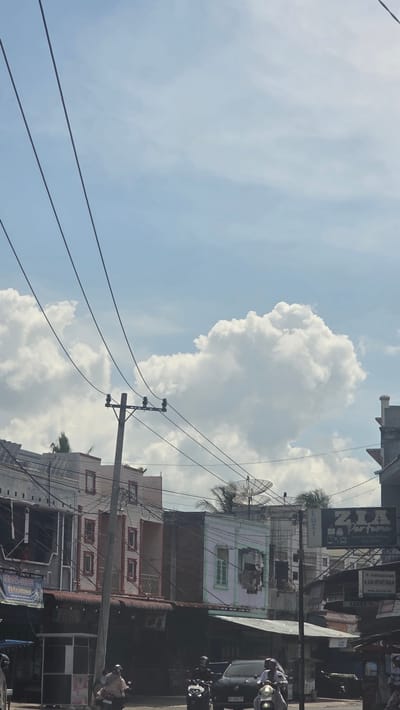 The image captures a street scene in Teungoh Baro, Indonesia, under a partly cloudy sky. Buildings line the street, displaying diverse architectural styles and signage, including commercial advertisements. Overhead power lines crisscross the view, connected to utility poles. The presence of satellite dishes indicates access to broadcast television services. Motorbikes with riders are visible on the street, suggesting the area is moderately active with vehicular traffic. The lighting indicates daylight hours. The composition of the shot is angled upwards, prioritizing the sky and clouds relative to the buildings and street below.