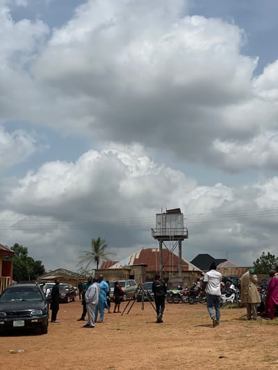 In Kassa, Nigeria, under a cloudy sky, a group of people are gathered in an open, dirt lot. Several vehicles are parked, including a black sedan in the foreground with a license plate that reads "PKN 631 KM". To the right of the sedan, a group of men are engaged in conversation, some dressed in traditional attire. Further back, a water tower stands tall, overlooking the scene. Motorcycles are also visible, parked together, suggesting a mode of transportation for some of the attendees. The background reveals residential buildings with corrugated metal roofs, indicative of the local architecture. The overall atmosphere appears to be a community gathering or event.