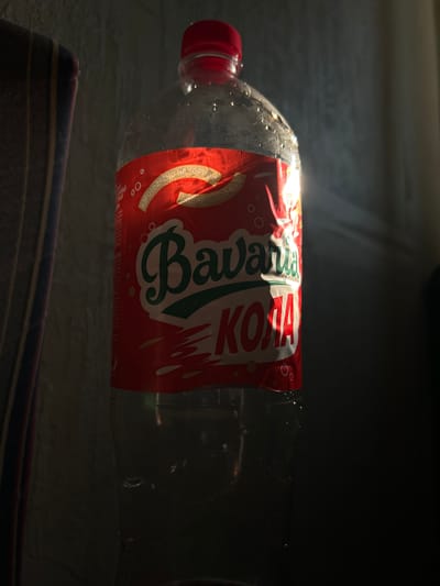 A clear plastic bottle with a red cap and a red label is positioned vertically against a textured, light-colored wall. The label features the brand name "Bavaria" in green script and "КОЛА" in red block letters. The bottle is partially filled with a dark liquid, and condensation droplets are visible on its surface, particularly on the upper portion. Sunlight is strongly illuminating the right side of the bottle, casting a bright highlight and creating a shadow on the wall behind it. To the left of the bottle, a striped fabric object, possibly a towel or curtain, is visible, also partly illuminated by the sunlight. The overall lighting suggests a bright, natural light source. The scene appears to be indoors, likely in a domestic setting. The city of Beslan, Russia, is not discernable from the visual elements within the image itself; its inclusion in a description would require external information.