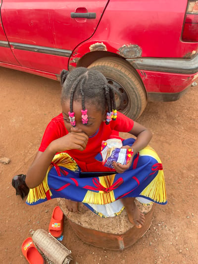 A young girl with braided hair sits on a tree stump on a dirt ground in Otukpa, Nigeria. She wears a red t-shirt and a brightly colored dress in shades of yellow, blue, and red. Pink heart-shaped clips adorn her hair. She holds a snack in her hand, looking downward. Nearby are a pair of orange sandals and a white purse with a chain. A red car with visible bodywork and a visible tire is partially in the shot.