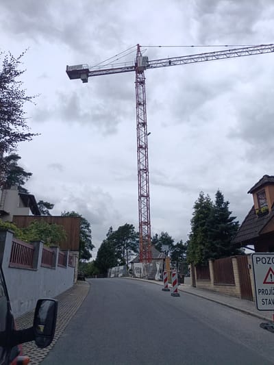 This is a wide shot of a residential street in Doksy, Czechia, on an overcast day. Dominating the scene is a large red construction crane, its jib extending horizontally across the sky. The street slopes upwards and curves to the right, lined with gray and brown fences, trees, and houses. To the right, a warning sign with a triangle and exclamation mark, along with text in Czech, indicates a hazard. Construction barriers, including red and white striped traffic pylons, are also visible on the side of the road, suggesting ongoing construction. The environment is primarily residential, with a mix of mature trees and the architectural styles of local housing.