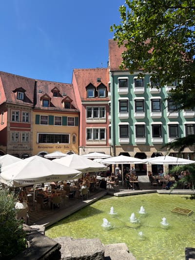 This image depicts a sunny outdoor seating area of a restaurant called "Restaurant Cafe Zeis" in Bamberg, Germany. The restaurant is situated in a square with buildings characterized by their traditional German architecture, featuring red tile roofs and colorful facades, including a prominent yellow building with green shutters. White umbrellas provide shade for tables occupied by patrons enjoying their meals and drinks. In the foreground, a fountain with multiple jets sprays water into a bright green pool, reflecting the clear blue sky. Several people are visible seated at tables, engaged in conversation or dining. Lush green trees and foliage frame the scene on the right side of the image. The ground is paved with interlocking bricks.