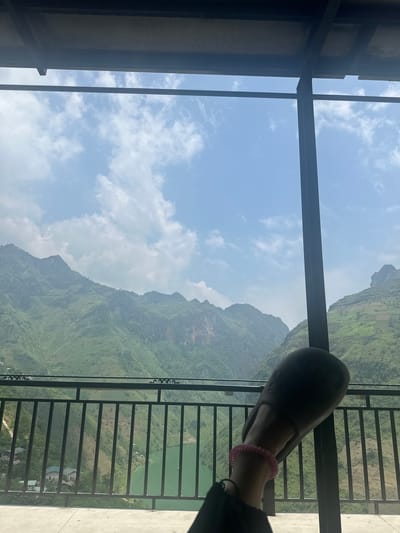 The image presents a view from a structure with dark metal framing and railings. Outside, there's a mountainous landscape, predominantly green with vegetation. A body of water, also green, is visible behind the railings. A part of a person's leg and foot, wearing a sandal and a pink scrunchie-like accessory, is in the foreground, partially obscuring the view. The sky is blue with scattered clouds. The location is likely Mèo Vạc Commune, Vietnam, based on the characteristic mountainous terrain.