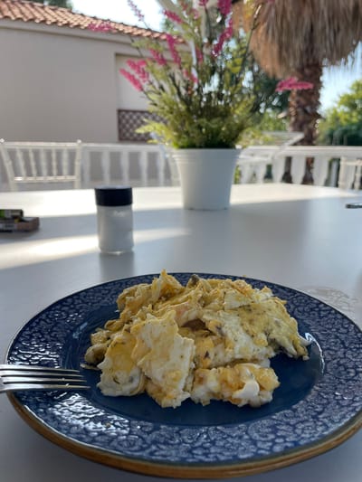 An outdoor dining scene is depicted in Skala Avdiron, Greece, with a plate of scrambled eggs as the central focus. The eggs are served on a dark blue patterned plate, positioned on a white table. A fork rests to the left of the plate. In the background, out of focus, a salt shaker, a pack of cigarettes, and a white flower pot containing pink flowers are visible. Beyond that, a white railing and a palm tree suggest a resort or beachfront setting. The lighting indicates it is daytime.