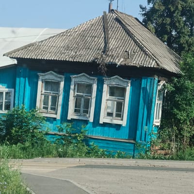 The image depicts a weathered, bright blue wooden house with a corrugated roof, situated in the city of Kambarka, Russia. The house has several windows with ornate white wooden trim, and is partially obscured by lush green foliage. A paved road and a pedestrian crossing are visible in the foreground. The sky is clear and blue. There are no people or animals visible in the frame.