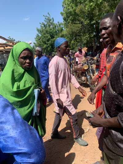 This media file depicts a scene in Boi A, Nigeria, with several people gathered outdoors under a bright sky, suggesting it is daytime. The group appears to be a mix of adults, some wearing traditional Islamic attire, including headscarves and caps. One individual, in the foreground and prominently featured, wears a bright green headscarf and is looking upwards.

The subjects are positioned in what seems to be a dusty, outdoor environment, with hints of trees and what might be rudimentary structures in the background. There are motorcycles present, indicating a mode of transportation or possibly a marketplace setting.

One notable detail is a person holding a wad of cash, possibly engaged in a transaction or distribution. The overall atmosphere suggests a communal gathering, with individuals appearing engaged in conversation or observation. The lighting and shadows indicate a sunny day. The people's facial expressions are largely neutral or contemplative, though it's difficult to discern specific emotions due to the image's orientation and quality. The presence of multiple individuals in traditional clothing and the rural setting are strong location cues for Boi A, Nigeria. No legible text is visible in the media file.