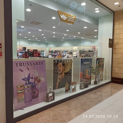 The image shows the storefront of "ESTASI profumerie" in Modugno, Italy. The store is a perfume shop with a large glass display window. Inside, shelves are stocked with various perfumes and beauty products from brands like L'Oreal, Dior, and Pupa. The display window features several large posters advertising different perfume brands, including Trussardi, Azzaro, Rabanne, and Dolce & Gabbana. One poster shows a man with the text "AZZARO WANTED" and another features a woman with "DOLCE & GABBANA Light Blue." The floor is tiled with a mosaic pattern. A date and time stamp "24.07.2026 10:24" is visible in the lower right corner of the image, suggesting this image was taken on July 24, 2026, at 10:24 AM. There are no people clearly visible inside or outside the store. The overall impression is that of a well-lit, modern retail space.