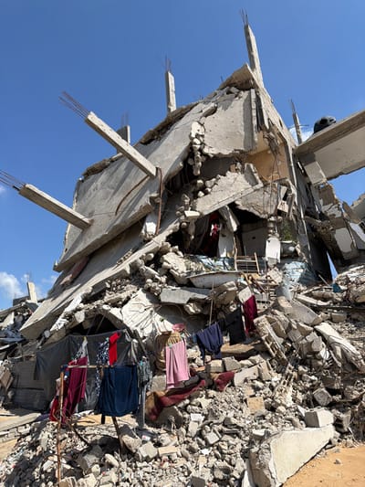 The image displays the severely damaged remains of a multi-story reinforced concrete building in Jabalia, Palestinian Territory, beneath a clear blue sky. The structure exhibits extensive collapse, with concrete slabs and pillars fractured and dislodged at acute angles, exposing twisted rebar. A large volume of pulverized concrete and structural debris forms a substantial rubble pile dominating the foreground and base of the damaged building. In the lower-center, a clothesline is suspended over the rubble, laden with drying garments including dark blue, red, pink, and patterned fabrics. Behind the clothesline, a dark tarp or fabric is visible, attached to the compromised lower section of the building. A white bathtub is observable among the debris near the building's foundation. The overall scene indicates comprehensive structural destruction with evidence of makeshift human adaptation within the immediate environment.