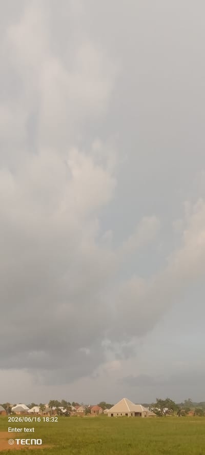 The image depicts a wide shot of a rural or suburban landscape, possibly in or around Gumel, Nigeria. The sky is heavily overcast with gray and white clouds, suggesting a potential for rain or a dull day. In the lower third of the image, a flat, open field of green grass stretches towards a cluster of houses. These houses appear to be in various stages of construction, with some showing visible framing and unfinished exteriors. One house in particular, on the right side of the frame, is a prominent, lighter-colored structure with a distinct pyramidal roof. The scene is devoid of people or any apparent activity. Text overlays in the lower left corner indicate a date of "2026/06/16 18:32" and a "TECNO" watermark, suggesting this is a photograph taken with a Tecno mobile device. The overall impression is one of a quiet, developing area under a cloudy sky.