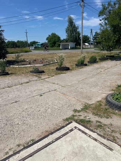 A person is cycling on a road in Malyi Trostianets, Ukraine. The road is paved and a few buildings are visible in the background, along with trees and a power line pole. There is a flowerbed in the foreground, decorated with flowers and some tires used as planters. The sky is blue with scattered clouds.