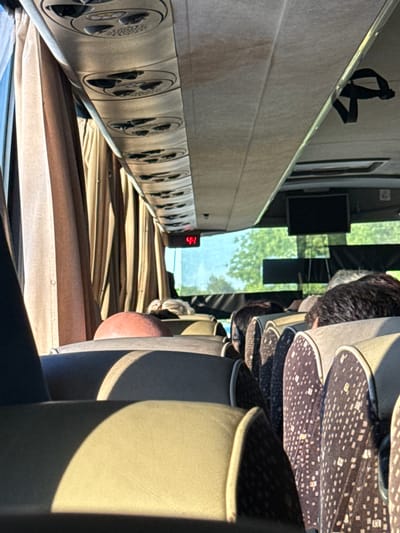 The image depicts the interior of a bus, viewed from the perspective of a passenger seated towards the rear. The ceiling of the bus is visible, featuring circular air vents and fabric upholstery. Curtains are drawn along the windows, casting a warm, diffused light into the cabin.

In the foreground, rows of empty bus seats with patterned upholstery are visible, bathed in sunlight. Some seats in the middle ground are occupied by passengers, whose heads and shoulders are visible. One passenger in the middle ground has a head of dark hair and another appears to have lighter colored hair, partially obscured.

A red digital display showing the number "44" is visible near the front of the bus, above the passengers. Further ahead, a dark rectangular screen, possibly a monitor, is mounted on the ceiling. Outside the windows, greenery and trees are visible, suggesting the bus is traveling through a rural or suburban area.

The location is specified as Izvorovo, Bulgaria, though the image itself provides no direct visual evidence to confirm this. The overall impression is one of a daytime journey on a public transport vehicle.