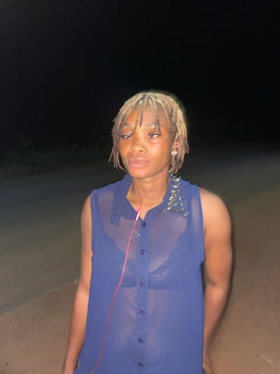 A person with blonde dreadlocks and wearing a sheer blue top is standing outdoors. The background is dark, suggesting night. The ground is dirt. The location is Ogbe, Nigeria. The person is wearing pink headphones. The top has studs on the shoulder.