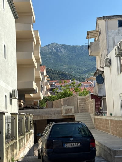 The media file depicts a narrow, uphill street in Lapčići, Montenegro, on a sunny day. The scene is dominated by buildings and a dark blue Volkswagen Passat van parked on the right side of the street.

The buildings are multi-story, with a mix of architectural styles. On the left, a light-colored building features geometric, stepped balconies. Further up the street, more buildings with terracotta roofs and white facades are visible, interspersed with trees. A large, rocky, forested mountain rises in the background, creating a dramatic backdrop.

The Passat van has a visible license plate that reads "CT AA661". Part of the van's interior is visible through the rear window, including the word "PASSAT" on the glass.

The activity appears to be a static scene, with no people or overt actions taking place. The bright sunlight and clear blue sky suggest it is daytime, likely late morning or early afternoon. The presence of air conditioning units on some buildings and the general appearance of the architecture are typical of Mediterranean coastal towns. The overall impression is one of a quiet, residential street in a hilly, scenic location.