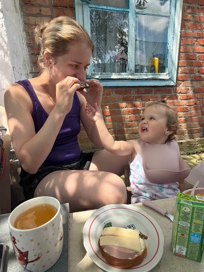 In Komsomolskiy, Russia, on a sunny day, a woman with blonde hair tied in a bun, wearing a purple tank top and dark shorts, sits outdoors and eats. She is seated next to a toddler, who wears a white onesie with bunny prints and a pink bib. The toddler is looking up and to the right, with their mouth open. In front of them, on a table covered with a beige tablecloth, are a mug of amber-colored liquid, a plate with a sandwich topped with cheese and ham, a pink-handled knife, and a juice box. The background shows a brick wall and a blue-trimmed window with a lace curtain. The scene suggests a casual meal in an outdoor setting, possibly a backyard or patio.