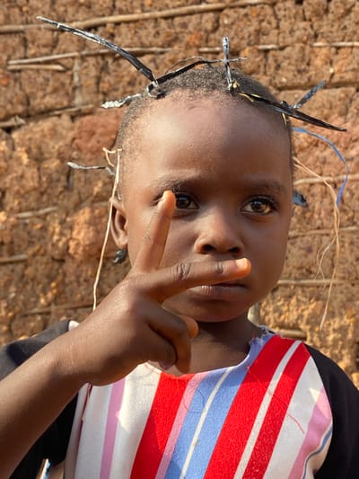 A young child of African descent stands centered in the frame, facing the camera directly. The child has dark skin, short dark hair, and brown eyes. Their head is adorned with several thin, dark, metallic-looking objects, appearing to be attached to the hair. The child's right hand is raised, with the index and middle fingers extended and touching their lips, as if in thought or to quiet themselves. The child is wearing a shirt with a black base and a brightly colored striped pattern of red, white, blue, and pink across the chest. The background consists of a textured, reddish-brown wall, possibly made of mud or brick, with visible horizontal and vertical elements. The lighting suggests it is daytime, casting a slight shadow on the child's face. The overall impression is a portrait captured outdoors, with a rustic environment suggested by the background. This image was captured in Ufu Esioni, Nigeria.