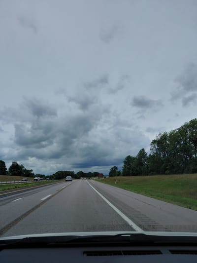 The image shows a roadside view from a vehicle traveling on a highway in Midway, United States of America. The sky is overcast with heavy, dark clouds, suggesting the possibility of rain or a storm. A line of trees borders the right side of the road, with a grassy area behind them. The highway itself is asphalt, with a white line dividing the lanes and a yellow line on the shoulder. Several cars are visible on the road, all traveling in the same direction as the camera. The overall mood is somewhat somber due to the cloudy weather, but the scene is otherwise unremarkable, depicting a typical travel day on an American highway. No text is visible in the image.