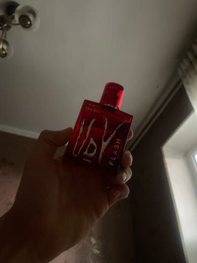 A hand holds a red glass bottle of "FLASH" perfume for men by "PARIS". The bottle is angled and illuminated from the left, casting shadows. The background is out of focus, showing a ceiling with a light fixture and a window with curtains. The image is taken from a low angle, looking upwards. The scene appears to be indoors, possibly in a residential setting.