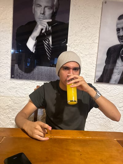 A medium shot depicts a young man drinking from a glass of orange juice. He is wearing a grey beanie and a dark t-shirt, and a silver watch is visible on his left wrist. He is sitting at a wooden table, and part of a black smartphone is on the table, near the right edge of the frame. Behind him, two framed black-and-white portraits are hanging on a white, textured wall. The location is presumed to be Chipilo de Francisco Javier Mina (Chipilo), Mexico.