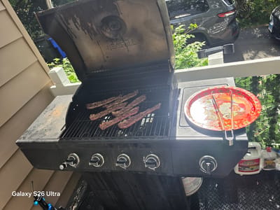 The image shows a Nexgrill propane barbecue with ribs on the grill. The barbecue is located on a wooden deck, with a beige vinyl-sided house on the left. In the background, there are two cars, green foliage, and blue garbage cans. A red decorative tray with tongs is on the side of the barbecue. Two bottles of cleaning solution and a metal can are on the ground next to the barbecue. A watermark for "Galaxy S26 Ultra" is visible in the bottom left corner of the image. The scene is set in West Hartford, United States of America.