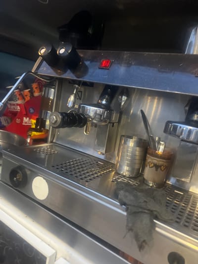 The image shows a close-up of an espresso machine, possibly located in Martil, Morocco, given the presence of "Martil" in a graphic below the machine. The machine is stainless steel and appears to be in use, with a towel partially covering the top. Coffee cups are situated on top. The machine has several controls and dispensers. The power switch is visible on the side.