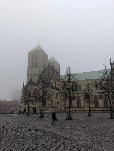 The image displays a foggy, overcast scene at Domplatz in Münster, Germany, with St. Paul's Cathedral (St. Paulus Dom) as the prominent subject. The large stone cathedral features two tall, square Romanesque towers topped with pyramidal, patinated green roofs and finials, their upper sections partially obscured by the dense fog. The main body of the cathedral exhibits elaborate Gothic architectural details, pointed arches, and numerous windows, with a distinct green-patinated roof stretching along its length.
In the foreground, the ground is covered with grey cobblestones, appearing damp. Several bare deciduous trees are symmetrically arranged in front of the cathedral, each supported by a dark metal tree guard. One individual, seen from behind, walks across the cobblestone square towards the cathedral, wearing a dark jacket, light blue jeans, and carrying a dark bag over their left shoulder. In the mid-ground to the left, several other indistinct figures are visible walking or standing near a multi-story building with a red-tiled roof, also partially shrouded in fog. A single streetlamp is visible among the trees to the left of the central figure. The overall lighting is diffuse and grey, indicative of the foggy conditions.