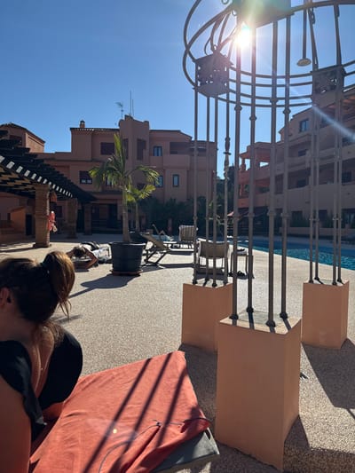 The image depicts an outdoor leisure area in Benahavís, Spain, under a clear blue sky with bright sunlight casting sharp shadows. The scene includes several terracotta-colored, multi-story buildings with windows and balconies, suggesting a residential or resort complex.
In the foreground, to the left, the back of a person with dark hair tied in a high ponytail is visible, wearing a dark top, positioned next to an orange towel draped over a grey surface. In the mid-ground, a person is lying face down on the textured, light-colored paved ground, appearing to be in a push-up or prone exercise position.
To the right, a large, ornamental metal structure, possibly a decorative shower or trellis, stands prominently with vertical bars and a curved top, casting long shadows. Beyond this structure, a blue swimming pool is visible. Several sun loungers are scattered around the pool area, one occupied by a reclining individual mostly obscured by the metal structure. Another sun lounger with a striped towel is visible unoccupied. A potted palm tree stands near the center of the image.
On the far left, a pergola with dark wooden beams and brick columns shades an area, where a red life preserver is hanging on a column. The overall environment is well-lit and appears well-maintained, indicating recreational use.