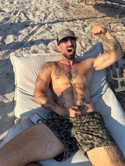 A man with extensive tattoos lies on a light gray cushioned lounge chair on a sandy beach. He is wearing a baseball cap and patterned shorts. The man is looking up and to his right with his mouth open, appearing to be shouting or laughing. His left arm is raised and flexed. Shadows from a nearby structure cast diagonal lines across the sand and chair, indicating it is daytime. The sand is light colored with scattered debris. A cell phone is visible on the cushion next to his leg. The setting is a beach, likely in a warm climate due to the man's attire. The overall mood is energetic and relaxed, suggesting a vacation or leisure activity.