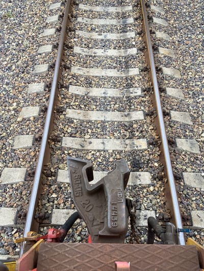 This is a high-angle, close-up shot of a railroad track in Mayachnoye, Russia. The tracks are made of steel rails supported by concrete sleepers, which are embedded in a bed of gravel. The scene is devoid of people. In the foreground, a section of a railway car is visible, with its coupling mechanism and associated hardware, including a heavy metal coupler, a black hose connected to a red valve, and a chain. The coupler has markings that appear to be in Cyrillic script and numerical identifiers: "60-06 20" and "ГоП 14439". The overall environment suggests an industrial or transportation setting.