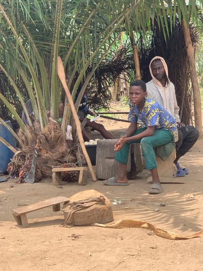 The image shows three men sitting near a palm tree, on the outskirts of Egbeda, Nigeria, located at coordinates 7.12 latitude, 5.362 longitude. The men are sitting on wooden platforms or logs. One man is wearing a white t-shirt and blue jeans, a second man is wearing a blue shirt with a floral print, and the third man is partially obscured by the palm tree, but appears to be wearing a light brown shirt. The men are talking and interacting with each other. There is a wooden paddle lying on the ground next to the palm tree. The environment is rural, and there is dry dirt and sand on the ground. The palm tree is in the foreground and provides a sense of shade for the men.