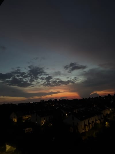 The image shows a dusky sky over a residential area, likely in Sindelfingen, Germany. The sky displays a gradient of colors, transitioning from a deep gray at the top to hues of orange and pink near the horizon. Several clouds are visible, some appearing dark and scattered, while others are more diffuse. Below the sky, the silhouettes of houses and trees form a dark landscape. A few lights are visible from within the houses, indicating that it is evening. The architecture of the houses suggests a suburban setting with row houses or similar dwelling types. The overall scene is tranquil and captures the transition from daylight to night.
