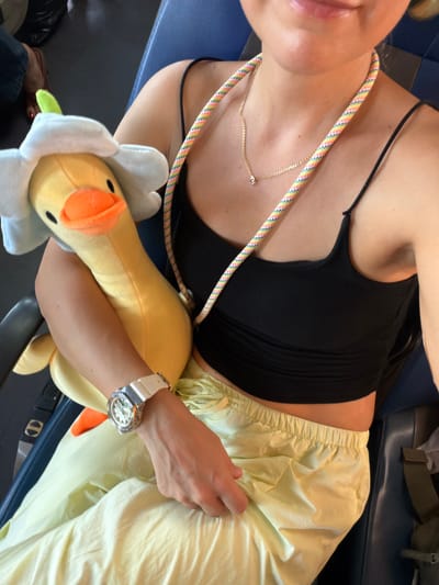 A young Caucasian woman is seated on a blue chair, holding a yellow duck plush toy wearing a white flower-shaped hat. She is wearing a black crop top, pale yellow drawstring pants, and a white watch with a diamond-encrusted bezel. A multicolored rope necklace hangs around her neck, and a thin gold chain with a small pendant rests below it. The woman's left arm is bent, cradling the duck toy, while her right hand is visible resting on her lap. The environment appears to be a public transportation setting, possibly an airplane or bus, with dark blue seating and a glimpse of a black bag in the background. The image is taken from a slightly elevated, personal perspective. There is no identifiable information in the image that places the scene in Hoa Xuan, Vietnam.