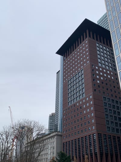 The image shows a tall building in Frankfurt am Main, Germany (latitude: 50.109, longitude: 8.673). The building is made of brick and has a large number of windows. It is located on the right side of the image, and a crane is visible on the left side of the image. The building is surrounded by other buildings, including a tall skyscraper with a glass facade on the left side of the image. There are trees on the ground in front of the building. The sky is overcast and gray. The image was taken during the day.