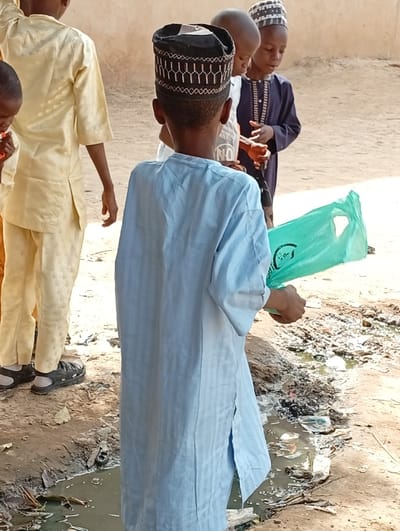This image captures a group of young boys in an outdoor setting in Fanisau, Nigeria, during what appears to be daytime under bright sunlight.
In the foreground, a boy stands with his back to the camera, wearing a light blue, long-sleeved tunic (jalabiya) and a black kufi cap adorned with intricate white embroidery. He holds a green plastic bag in his right hand. To his left, partially visible, another boy also faces away, dressed in a light yellow tunic and trousers, with black sandals on his feet.
Further back, two other boys are visible. One, facing right, wears a white shirt with the letters "NO" faintly visible on it, and another boy in a dark purple tunic and a striped cap is facing him. Their expressions are not clearly discernible, but they seem to be focused on something between them or interacting.
The ground is unpaved, appearing dusty and uneven, characteristic of a rural or semi-urban environment. A significant detail is the puddle of dirty, stagnant water in the lower part of the frame, filled with various debris including plastic items and leaves. This suggests a drainage issue or unkempt surroundings. The background features a light-colored wall or structure, possibly made of mud or concrete. The general attire of the boys, including the tunics and caps, indicates cultural or religious garments common in the region.